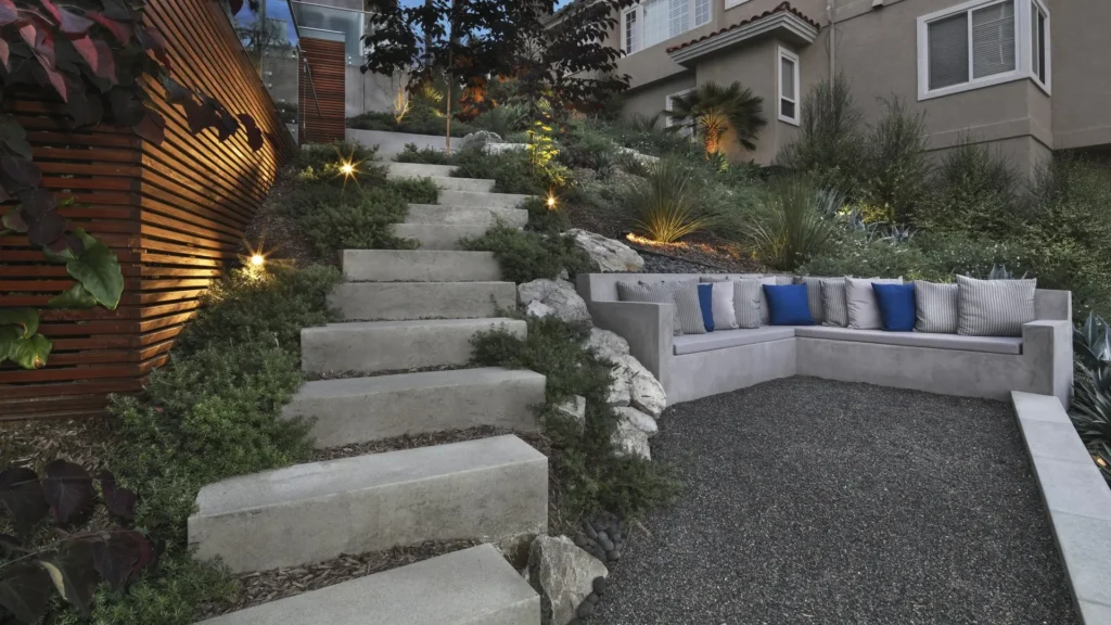 Modern hardscape design featuring stone steps leading to a cozy seating area with gray and blue cushions, surrounded by lush greenery and illuminated by soft lighting, showcasing Urban Landscape's expertise in landscape transformation in Orange County.