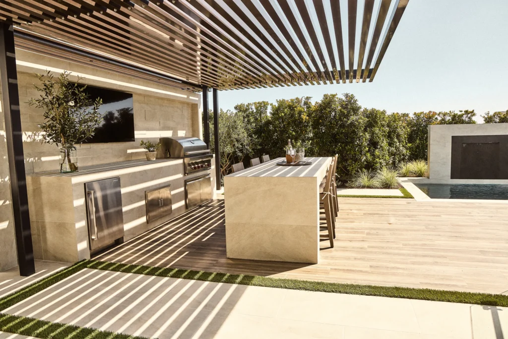 Outdoor kitchen with modern grill, sleek countertops, and dining area under a shaded pergola, surrounded by greenery and a pool, showcasing durable materials for outdoor living.