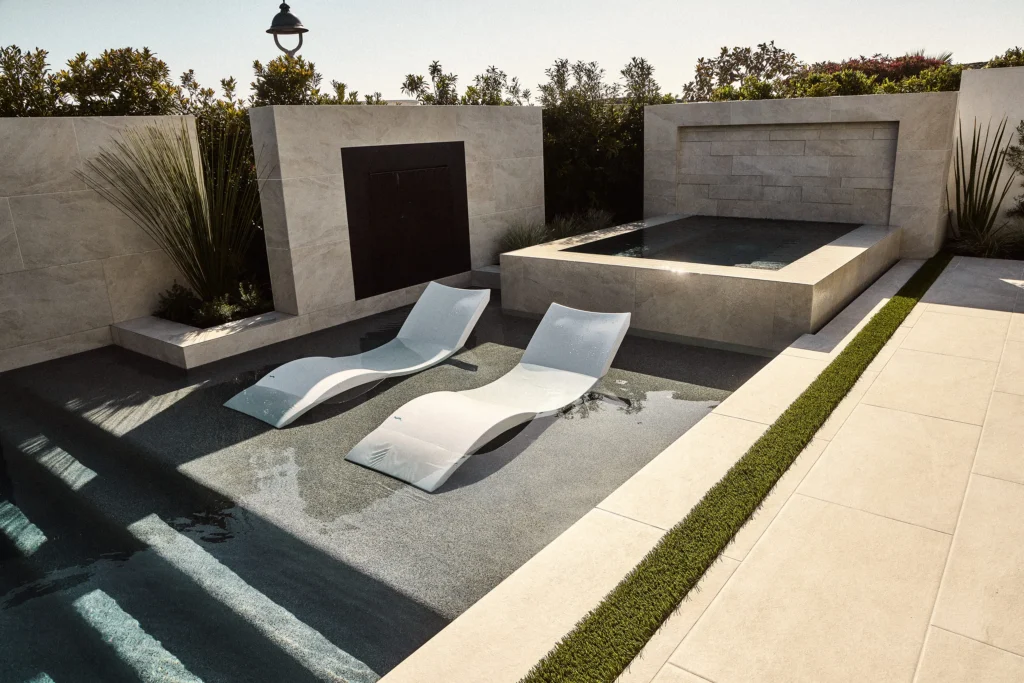 Luxury outdoor living area featuring modern lounge chairs beside a pool, surrounded by sleek stone walls and lush greenery, exemplifying high-end hardscape design for relaxation and comfort.